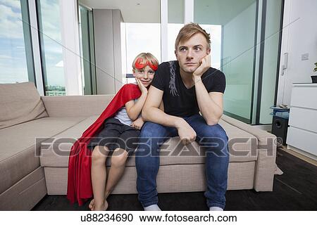 Stock Image - Portrait of smiling boy dressed in superhero costume sitting with sad father on sofa bed at home. Fotosearch
