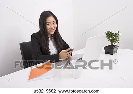 Stock Image - Portrait of smiling businesswoman using cell phone at office desk. Fotosearch