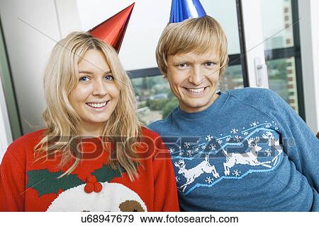 Stock Photo - Portrait of smiling couple wearing Christmas sweaters and party hats in living room at home. Fotosearch