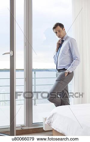 Portrait of worried businessman adjusting necktie by glass door in hotel View Large Photo Image Stock Photo - Portrait of worried businessman adjusting necktie by glass door in hotel. Fotosearch