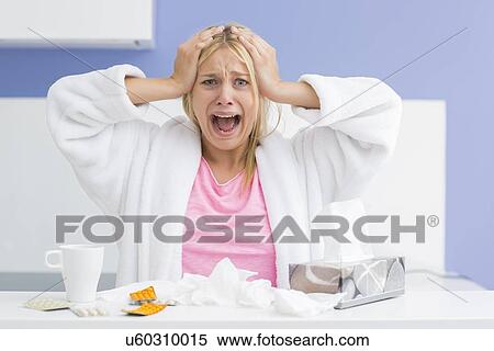 Stock Photography - Portrait of young exhausted woman screaming while suffering from headache and cold in kitchen. Fotosearch