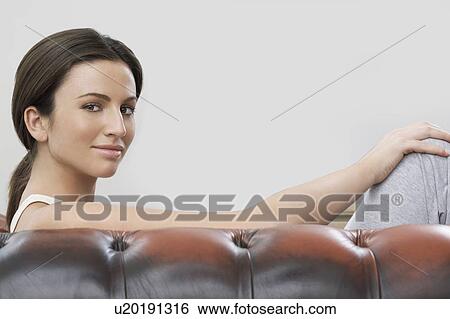 Stock Photograph - Portrait of young woman sitting on sofa against gray background. Fotosearch