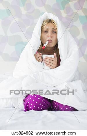 Stock Image - Portrait of young woman with coffee mug taking temperature while wrapped in quilt on bed. Fotosearch