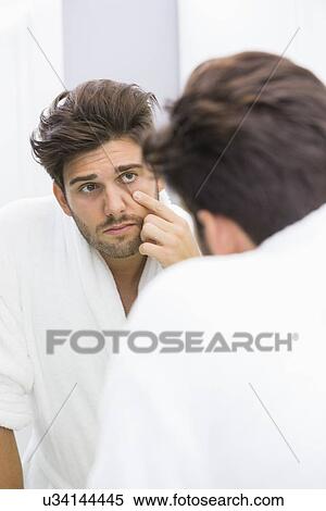 Stock Photography - Reflection of ill man examining eyes in mirror. Fotosearch
