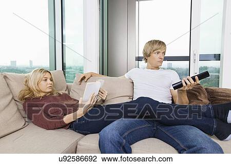 Stock Image - Relaxed couple reading book and watching TV in living room at home. Fotosearch