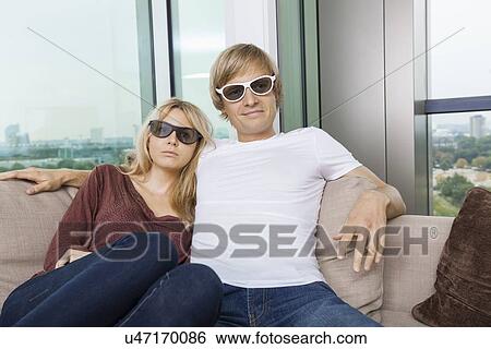 Relaxed couple wearing 3D glasses while sitting on sofa at home View Large Photo Image Stock Photograph - Relaxed couple wearing 3D glasses while sitting on sofa at home. Fotosearch