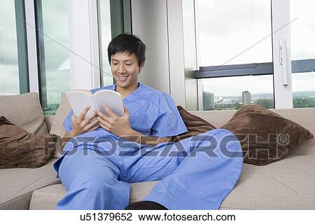 Stock Image - Relaxed mid adult man reading book in living room at home. Fotosearch