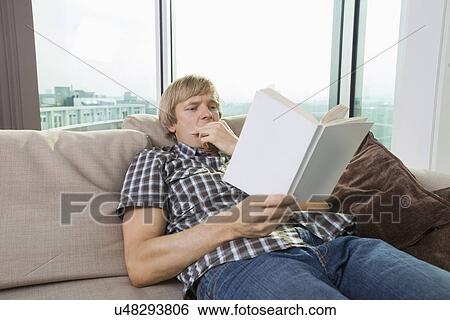 Stock Photograph - Relaxed mid-adult man reading book in living room at home. Fotosearch