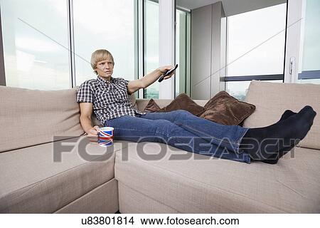 Picture - Relaxed mid-adult man watching television while having coffee on sofa at home. Fotosearch