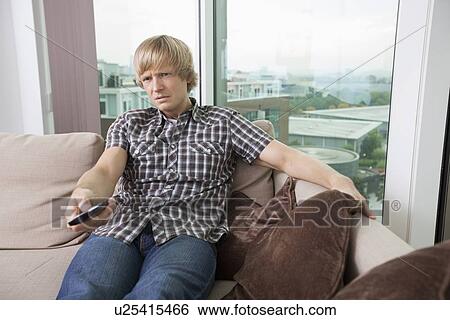 Stock Photograph - Relaxed mid-adult man watching television on sofa at home. Fotosearch