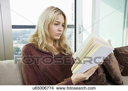 Picture - Relaxed young woman reading book in living room at home. Fotosearch