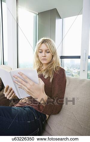 Stock Image - Relaxed young woman reading book in living room at home. Fotosearch