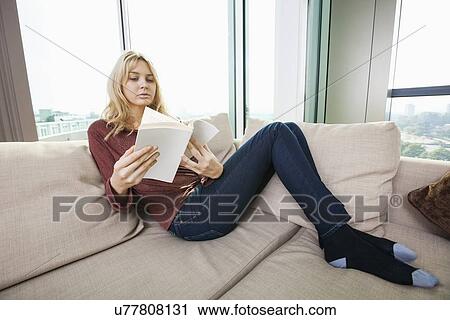 Stock Image - Relaxed young woman reading book in living room at home. Fotosearch