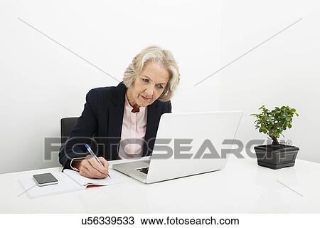 Senior businesswoman writing in book while using laptop at desk in office View Large Photo Image Stock Image - Senior businesswoman writing in book while using laptop at desk in office. Fotosearch