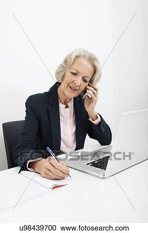 Senior businesswoman writing in book while answering cell phone at desk in office View Large Photo Image Stock Image - Senior businesswoman writing in book while answering cell phone at desk in office. Fotosearch