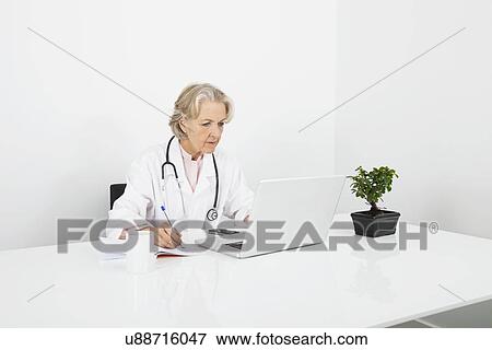 Stock Photo - Senior female doctor writing notes while looking at laptop in clinic. Fotosearch