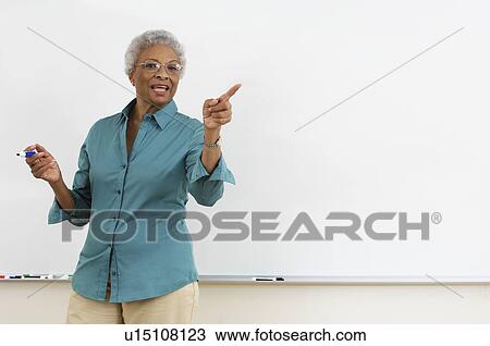 Senior teacher pointing while gesturing against white board in classroom View Large Photo Image Stock Image - Senior teacher pointing while gesturing against white board in classroom. Fotosearch