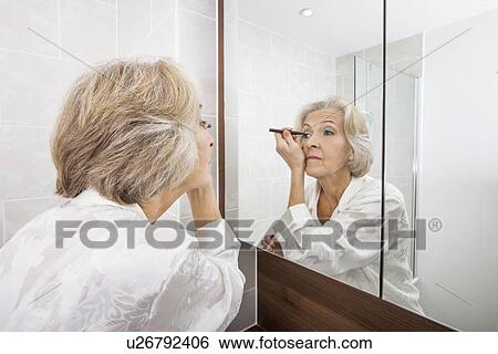 Stock Photograph - Senior woman applying eyeliner while looking at mirror in bathroom. Fotosearch