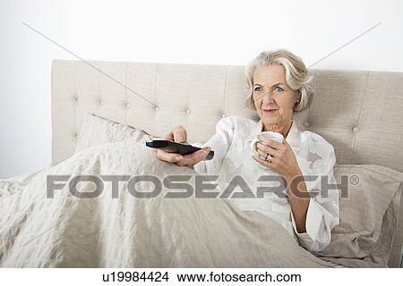 Senior woman watching TV while having coffee in bed View Large Photo Image Picture - Senior woman watching TV while having coffee in bed. Fotosearch