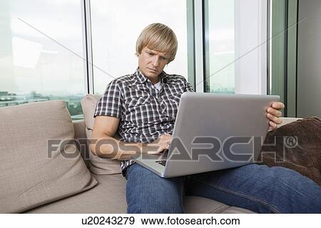 Serious mid-adult man using laptop on sofa at home View Large Photo Image Stock Photo - Serious mid-adult man using laptop on sofa at home. Fotosearch