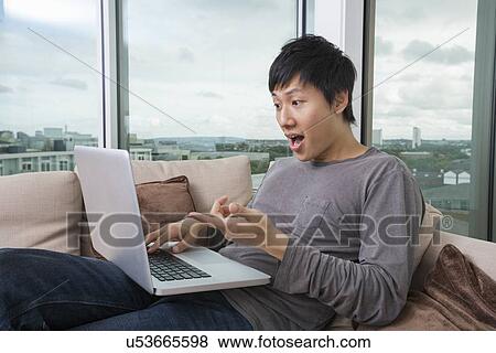 Stock Photo - Shocked man using laptop on sofa at home. Fotosearch