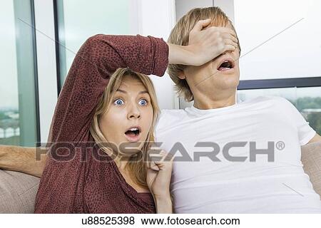 Stock Photo - Shocked woman covering man's eyes while watching TV at home. Fotosearch