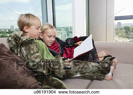 Picture - Siblings in dinosaur and vampire costumes reading picture book together on sofa bed at home. Fotosearch