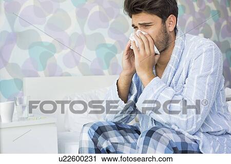 Stock Image - Sick man blowing his nose in tissue paper on bed at home. Fotosearch