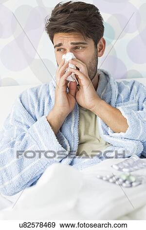 Stock Photo - Sick man blowing his nose in tissue paper on bed at home. Fotosearch
