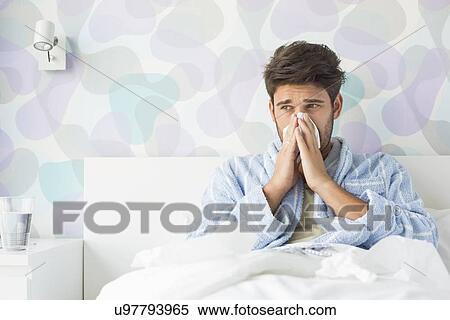 Stock Photography - Sick man blowing his nose while sitting on bed at home. Fotosearch