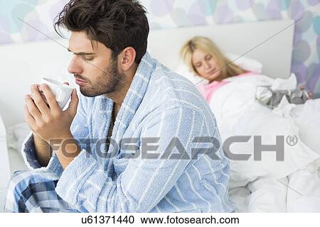 Stock Image - Sick man drinking coffee on bed while woman sleeping in background at home. Fotosearch