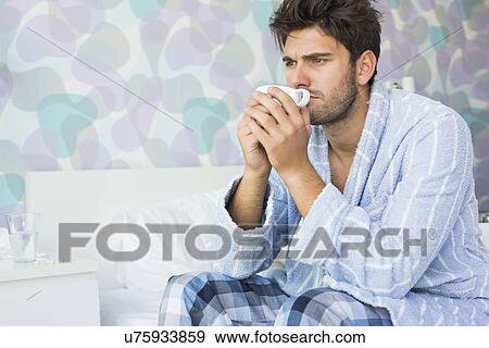 Sick man drinking coffee while sitting on bed at home View Large Photo Image Stock Photo - Sick man drinking coffee while sitting on bed at home. Fotosearch