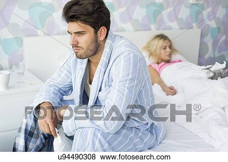 Stock Photo - Sick man sitting on bed with woman sleeping in background at home. Fotosearch