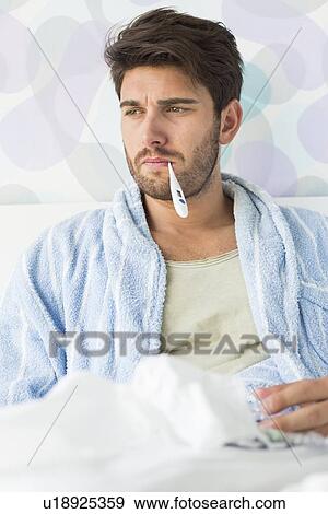 Stock Photo - Sick man with thermometer in mouth sitting on bed. Fotosearch