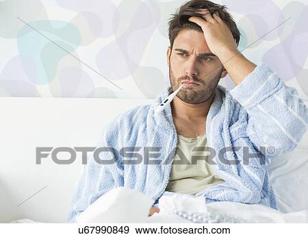 Stock Photo - Sick man with thermometer in mouth sitting on bed at home. Fotosearch