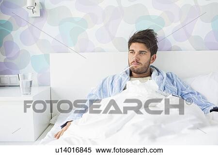 Stock Photography - Sick man with thermometer in mouth reclining on bed at home. Fotosearch