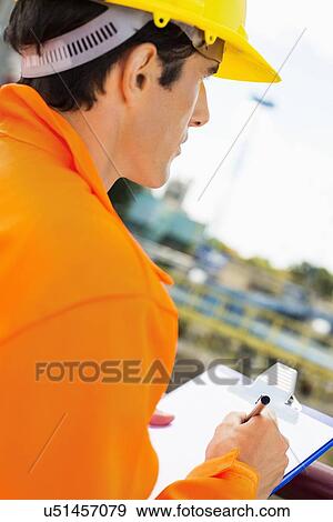 Stock Photo - Side view of construction worker writing on clipboard at construction site. Fotosearch