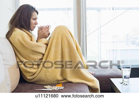 Stock Image - Side view of sick woman having coffee on sofa in living room. Fotosearch