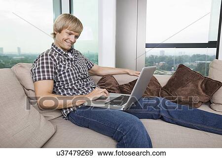 Stock Photo - Side view of smiling mid-adult man using laptop on sofa at home. Fotosearch