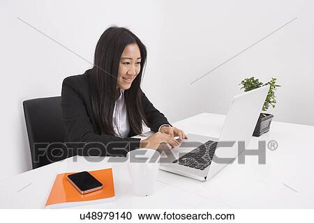 Stock Image - Smiling businesswoman working on laptop at office desk. Fotosearch