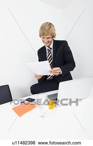 Smiling mid adult businessman reading document with laptop on desk at office View Large Photo Image Stock Photo - Smiling mid adult businessman reading document with laptop on desk at office. Fotosearch