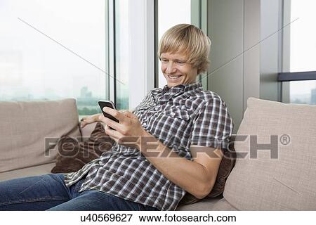 Smiling mid-adult man text messaging on sofa at home View Large Photo Image Stock Photo - Smiling mid-adult man text messaging on sofa at home. Fotosearch