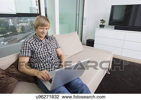 Stock Image - Smiling mid-adult man using laptop on sofa at home. Fotosearch