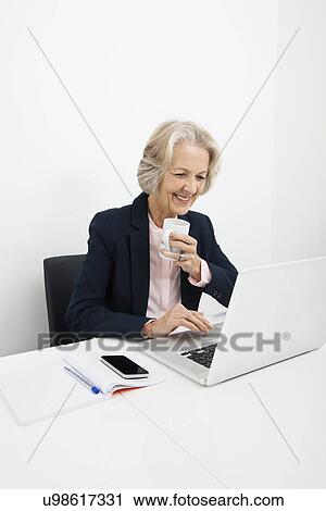 Smiling senior businesswoman having coffee while using laptop at desk in office View Large Photo Image Stock Image - Smiling senior businesswoman having coffee while using laptop at desk in office. Fotosearch