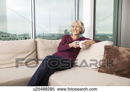 Stock Photography - Smiling senior woman with coffee mug relaxing on sofa at home. Fotosearch