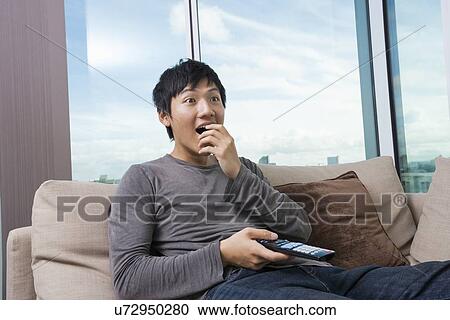 Stock Image - Surprised mid-adult man watching television on sofa at home. Fotosearch