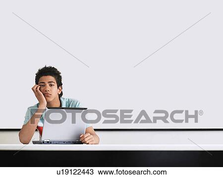 Stock Image - Teenage boy looking bored in front of whiteboard in classroom. Fotosearch