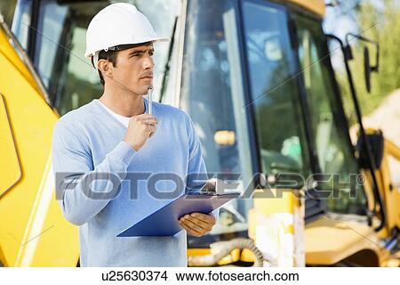 Picture - Thoughtful architect with clipboard and pen standing against earthmover at site. Fotosearch