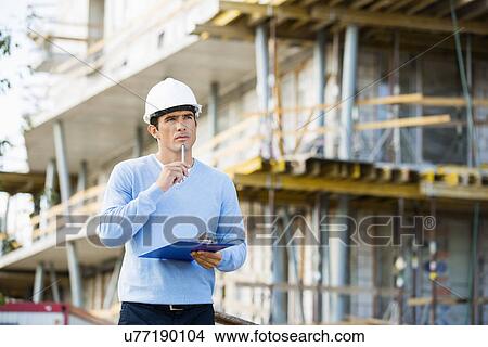 Picture - Thoughtful architect with clipboard and pen standing at site. Fotosearch