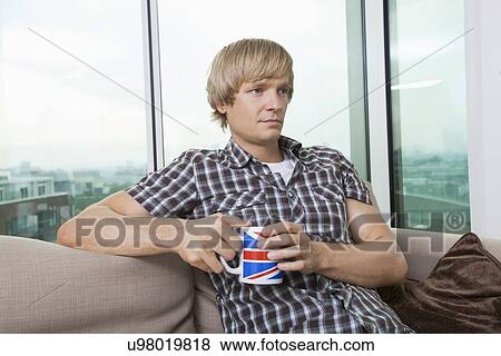 Thoughtful mid-adult man sitting with coffee cup in living room at home View Large Photo Image Stock Photo - Thoughtful mid-adult man sitting with coffee cup in living room at home. Fotosearch